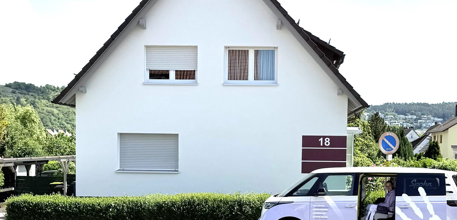 Frontansicht eines gepflegten Einfamilienhauses mit weißer Fassade und der Hausnummer 18; davor steht ein Firmenfahrzeug eines Malerbetriebs am Straßenrand.