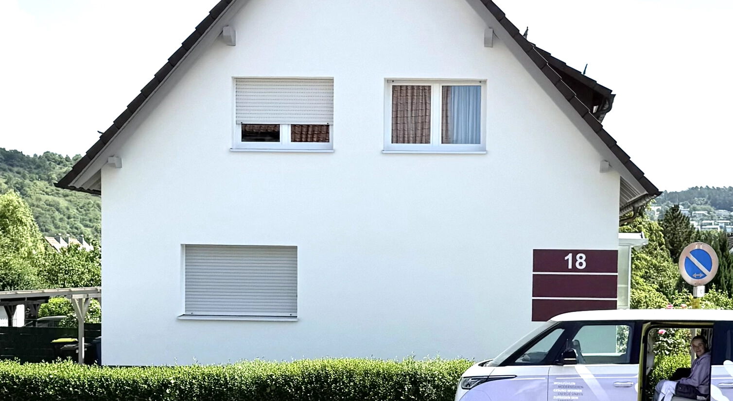 Frontansicht eines gepflegten Einfamilienhauses mit weißer Fassade und der Hausnummer 18; davor steht ein Firmenfahrzeug eines Malerbetriebs am Straßenrand.