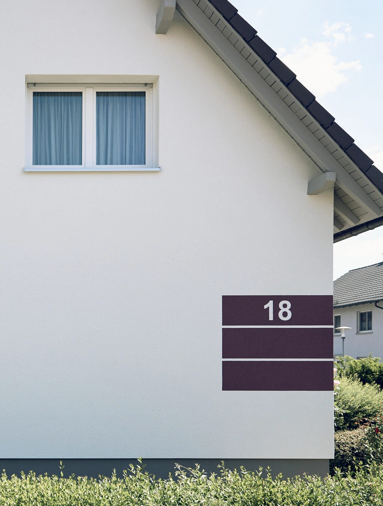 Seitenansicht eines Einfamilienhauses mit heller Fassade, kleinem Fenster im Obergeschoss und bordeauxfarbenem Fassadenelement mit der Hausnummer 18.