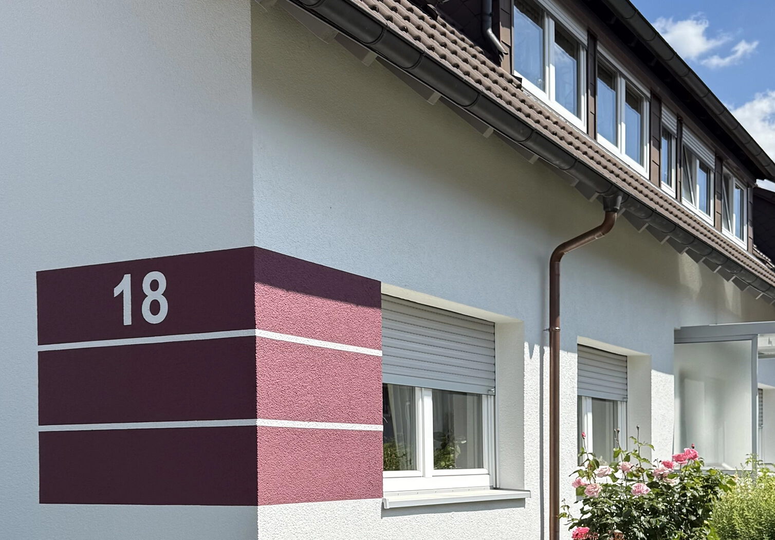 Außenansicht eines modern gestalteten Einfamilienhauses mit heller Fassade, bordeauxfarbenem Akzentstreifen und der Hausnummer 18, weißen Fenstern und blühenden Rosen im Vordergrund.