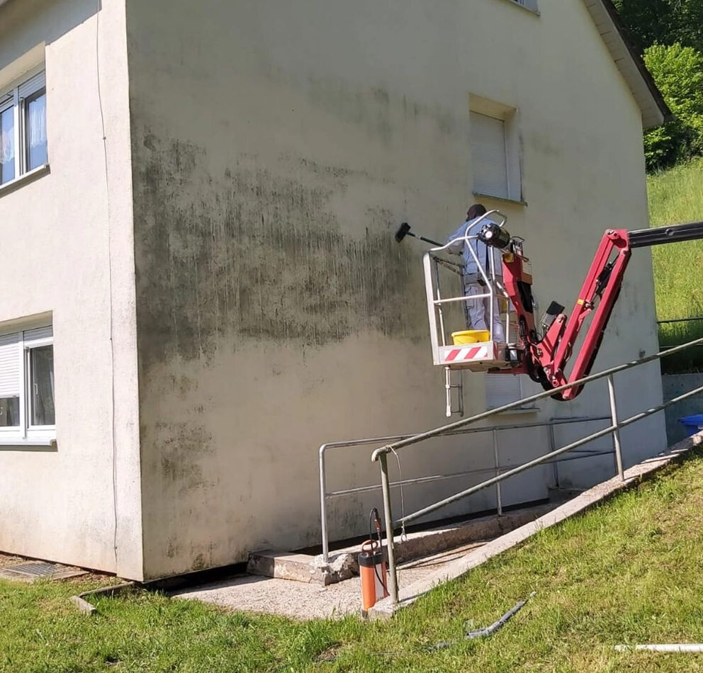 Fassadenreinigung eines Wohngebäudes: Mitarbeiter arbeitet mit Hebebühne an einer stark verschmutzten Putzfassade.