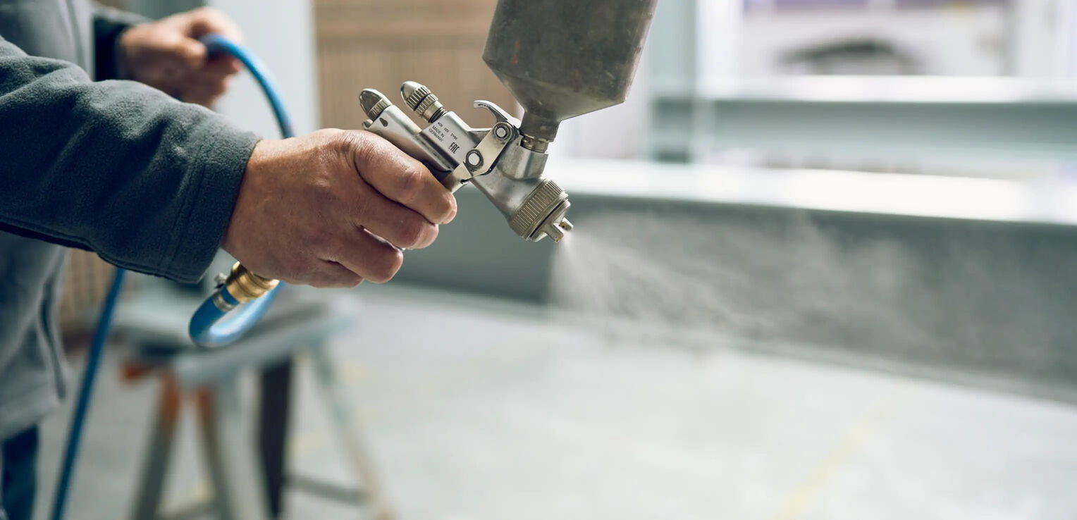 Eine Person hält eine Spritzpistole, aus der Nebel oder Farbe freigesetzt wird. Die Hand ist nah aufgenommen, der Hintergrund ist verschwommen. Ein blauer Schlauch ist an die Pistole angeschlossen.